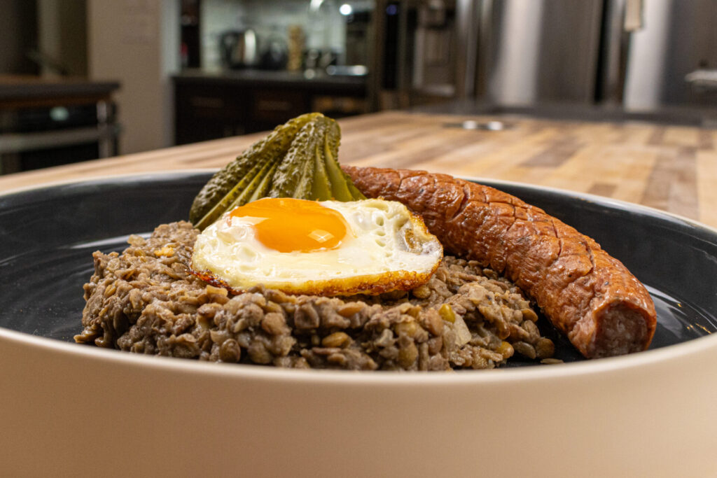 Lentil Meal on the black and white plate with cooked kielbasa, sunny side up egg and pickles.